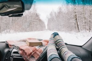 The inside view of a person's feet up on the dash with winter socks on. There is a winter blanket and a book inside the car as the view outside is snowy with snowflakes falling.