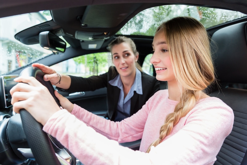A young teen blonde girl wearing a pink shirt. She is in the car with an instructor for her driving permit. 