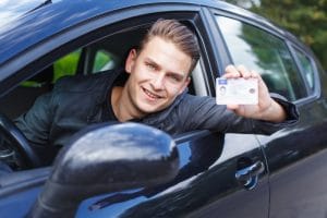 A young teen boy with braces that is hanging out of the driver's window holding a new driver's license.