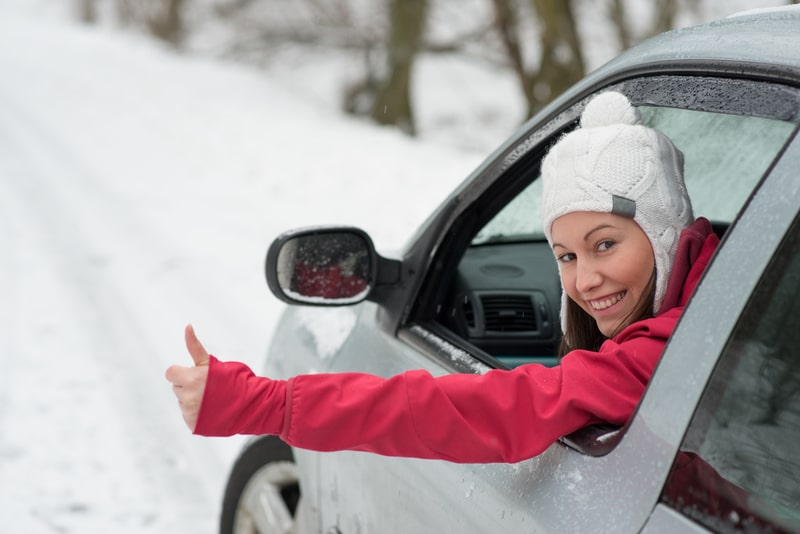 A woman that is about to drive in snow. Her window is rolled down and she is giving the camera a thumbs-up.