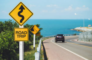 Sign that says "road trip" with a car in the background driving on a winding road.
